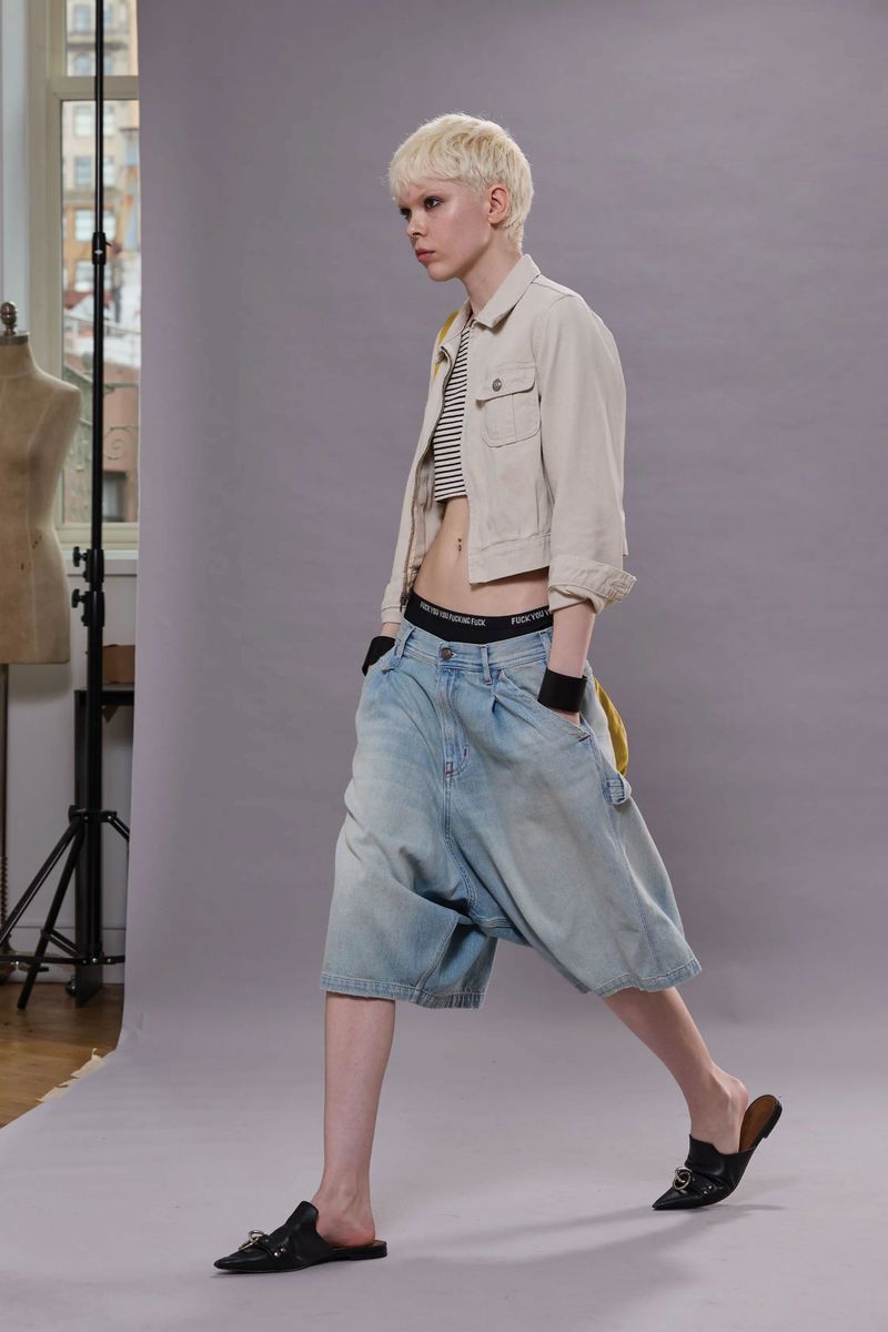 Fashion shoot: a model with pale skin and a blonde buzzcut strides confidently in oversized denim shorts and a neutral jacket.
