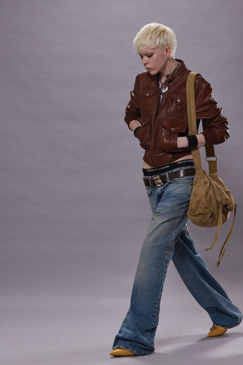 Young adult female in a brown leather jacket, jeans, and yellow shoes posing with a satchel against a gray studio background.