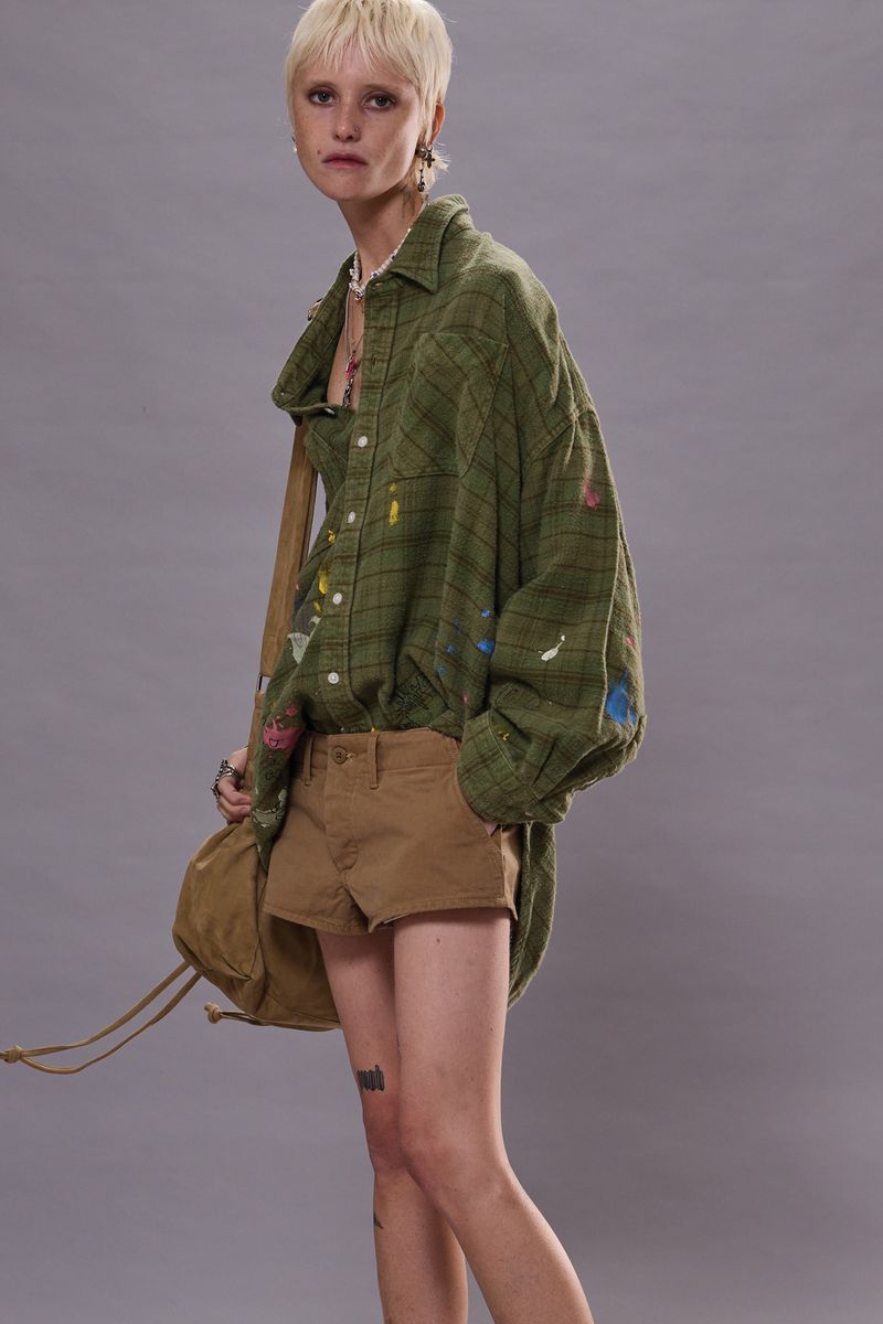 Adult woman with short blonde hair poses in a long green plaid shirt and brown shorts with a khaki-colored bag against gray.