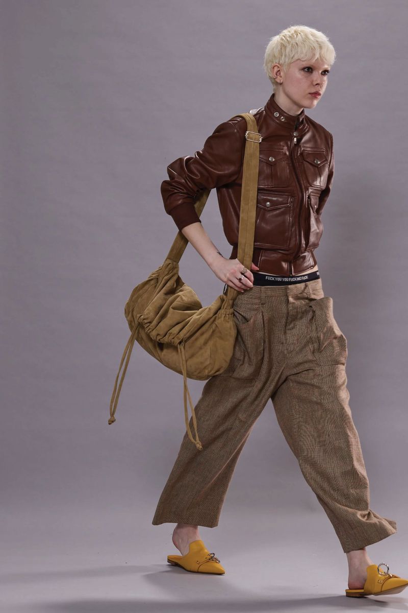 Fashion shot of a person with short blonde hair walking and wearing a brown leather jacket, loose brown pants, and a tan hobo bag.