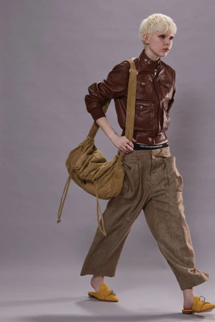 Fashion shot of a person with short blonde hair walking and wearing a brown leather jacket, loose brown pants, and a tan hobo bag.
