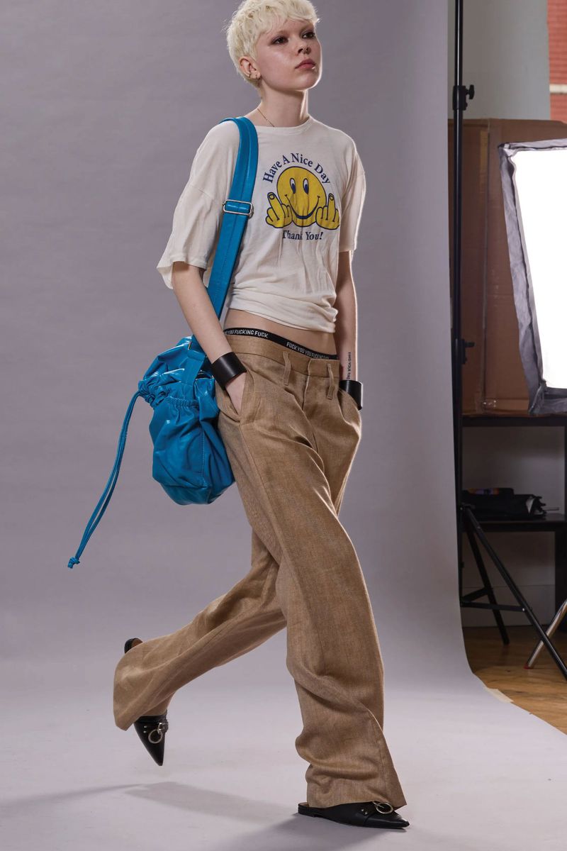 Adult female with blonde hair poses in a white tee shirt and tan trousers, carrying a blue shoulder bag in a studio environment.