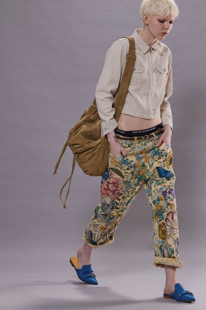 Woman in a cropped jacket, floral pants, and blue mules with a brown shoulder bag, standing indoors against a solid grey background.