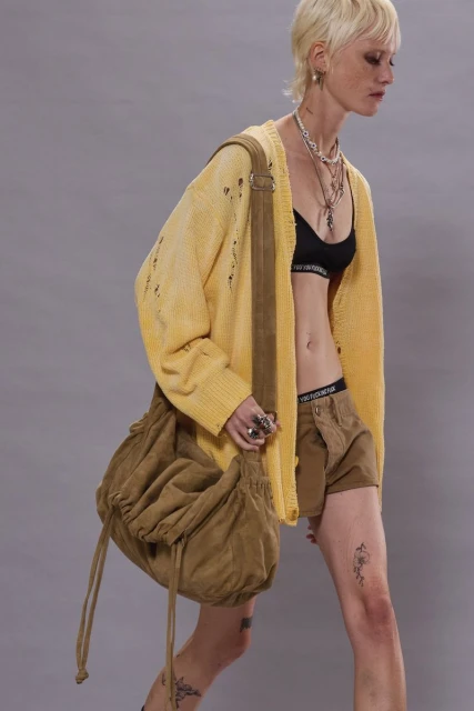 Adult female with blonde hair wearing a yellow cardigan, brown shorts, black bra, and a brown shoulder bag in a studio setting.