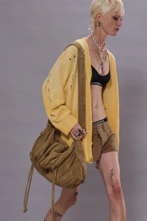 Adult female with blonde hair wearing a yellow cardigan, brown shorts, black bra, and a brown shoulder bag in a studio setting.