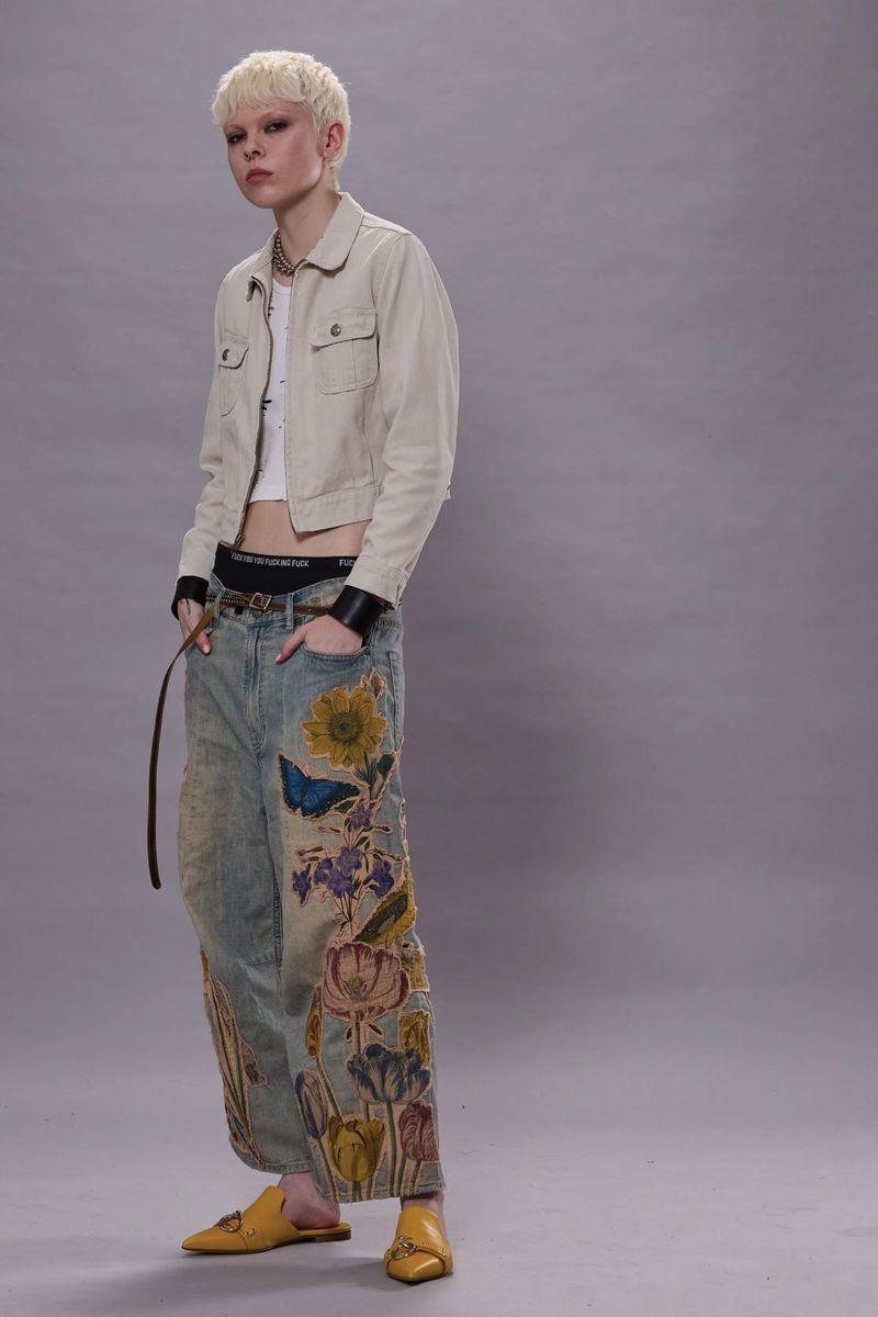 Model with blonde hair wearing a beige jacket and embroidered jeans against a grey studio backdrop.