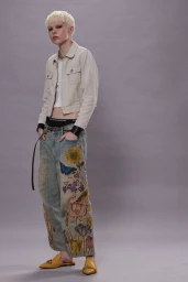 Model with blonde hair wearing a beige jacket and embroidered jeans against a grey studio backdrop.