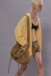 Model walking in a distressed yellow cardigan, black bralette, khaki shorts, and a slouchy crossbody bag against a grey backdrop.