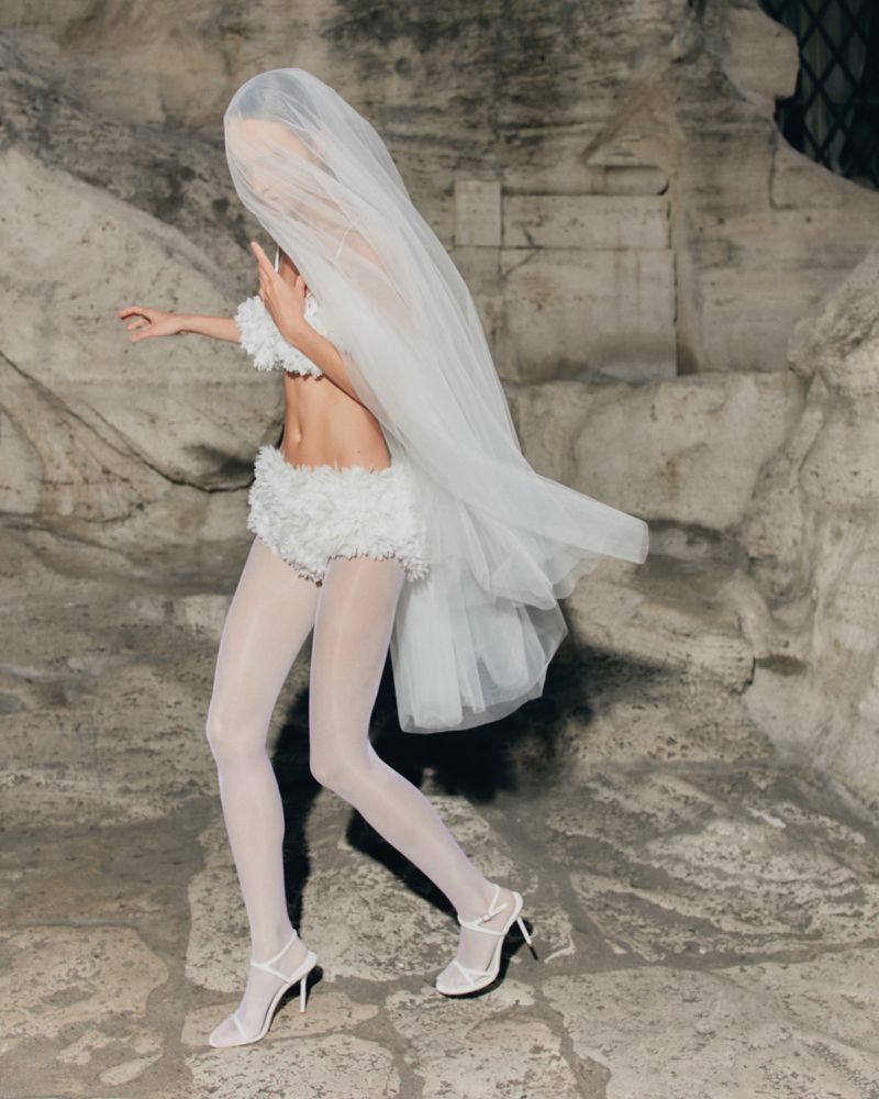Woman in white bra top, shorts, tights, and heels stands against an old stone wall with a large veil covering her head and body.