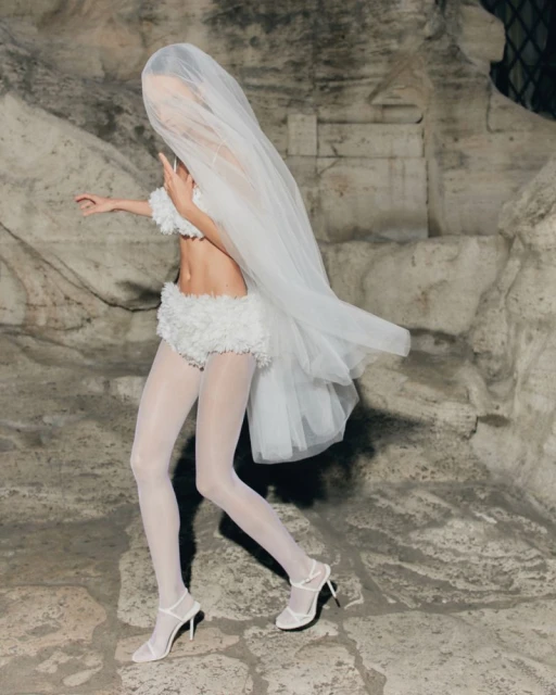 Woman in white bra top, shorts, tights, and heels stands against an old stone wall with a large veil covering her head and body.