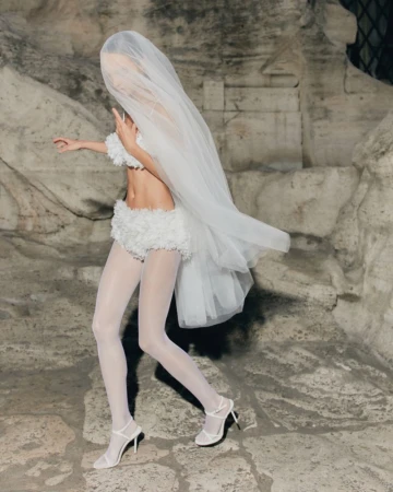 Woman in white bra top, shorts, tights, and heels stands against an old stone wall with a large veil covering her head and body.