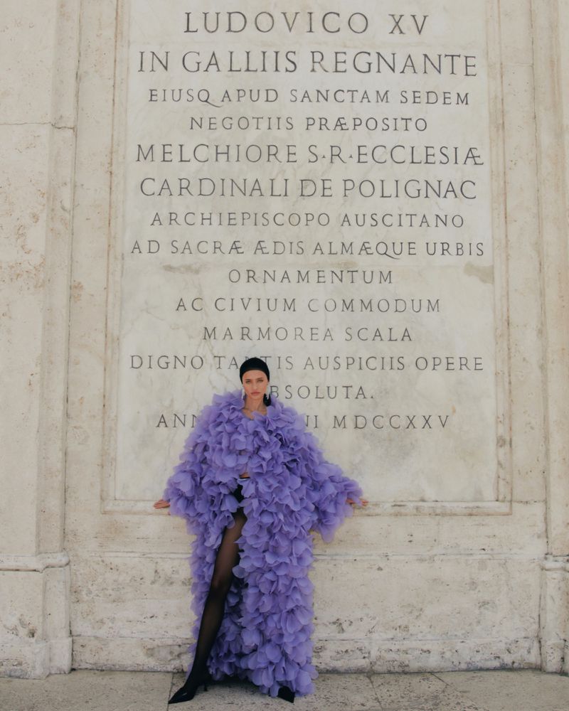 Woman in a lavender ruffled coat standing in front of an inscribed marble wall, wearing black tights and heels in an outdoor setting.