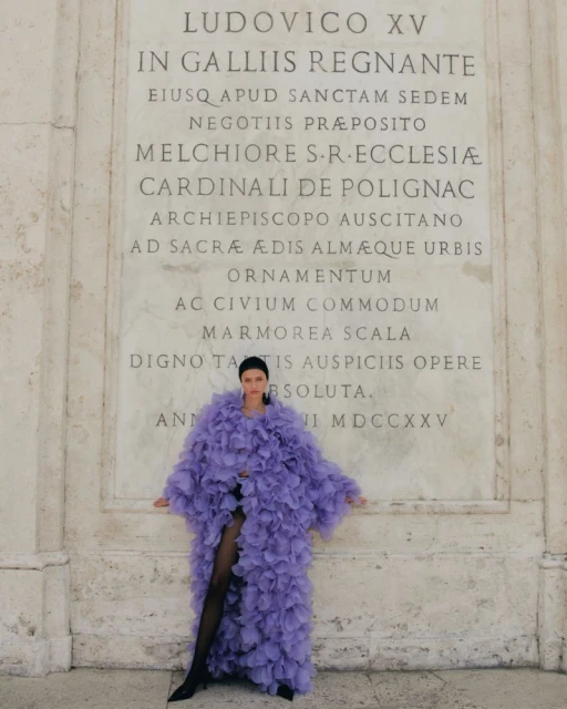 Woman in a lavender ruffled coat standing in front of an inscribed marble wall, wearing black tights and heels in an outdoor setting.