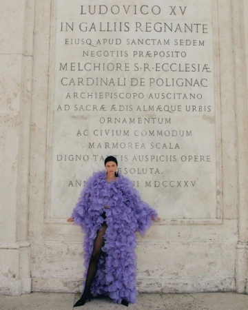 Woman in a lavender ruffled coat standing in front of an inscribed marble wall, wearing black tights and heels in an outdoor setting.