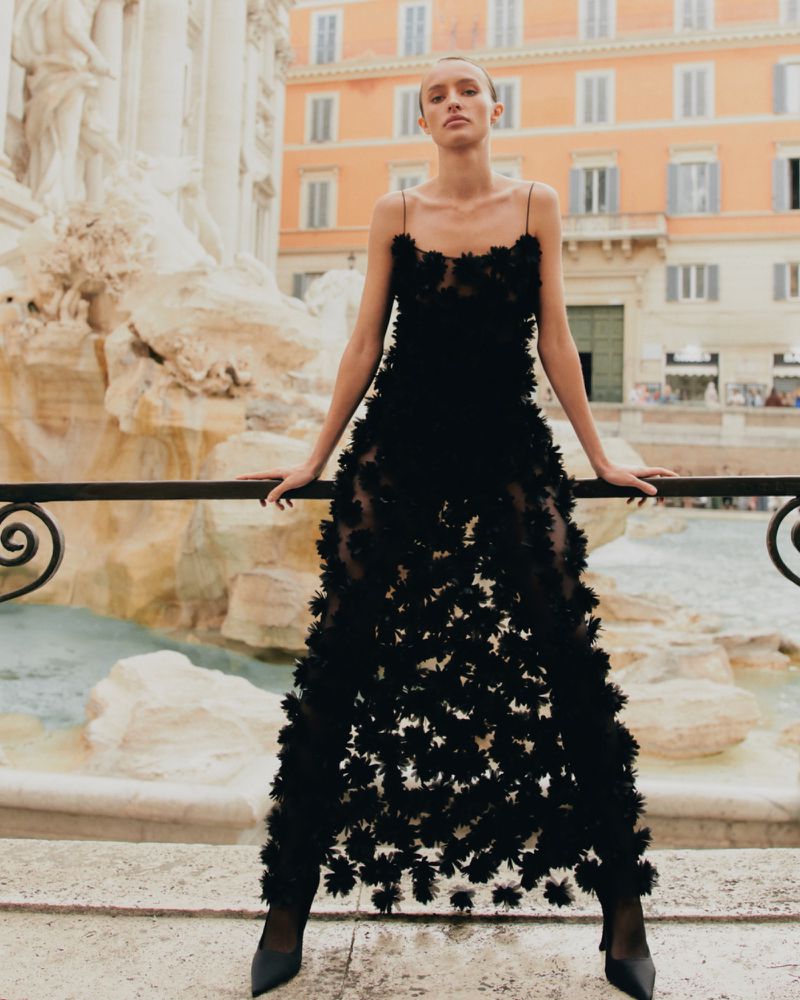 Woman in black floral dress poses in front of Trevi Fountain, Rome. She leans against a low railing, with buildings in the background.