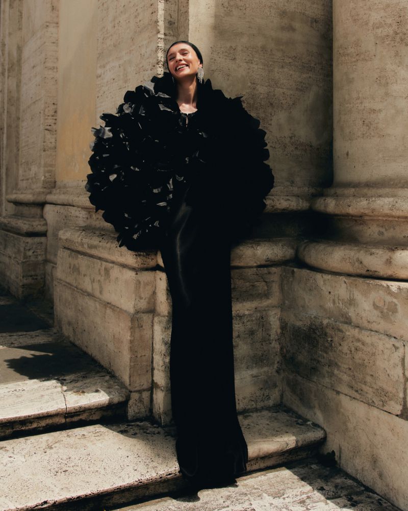 Elegant adult woman in black gown and ruffled cape posing on weathered stone steps near columns with a happy expression.
