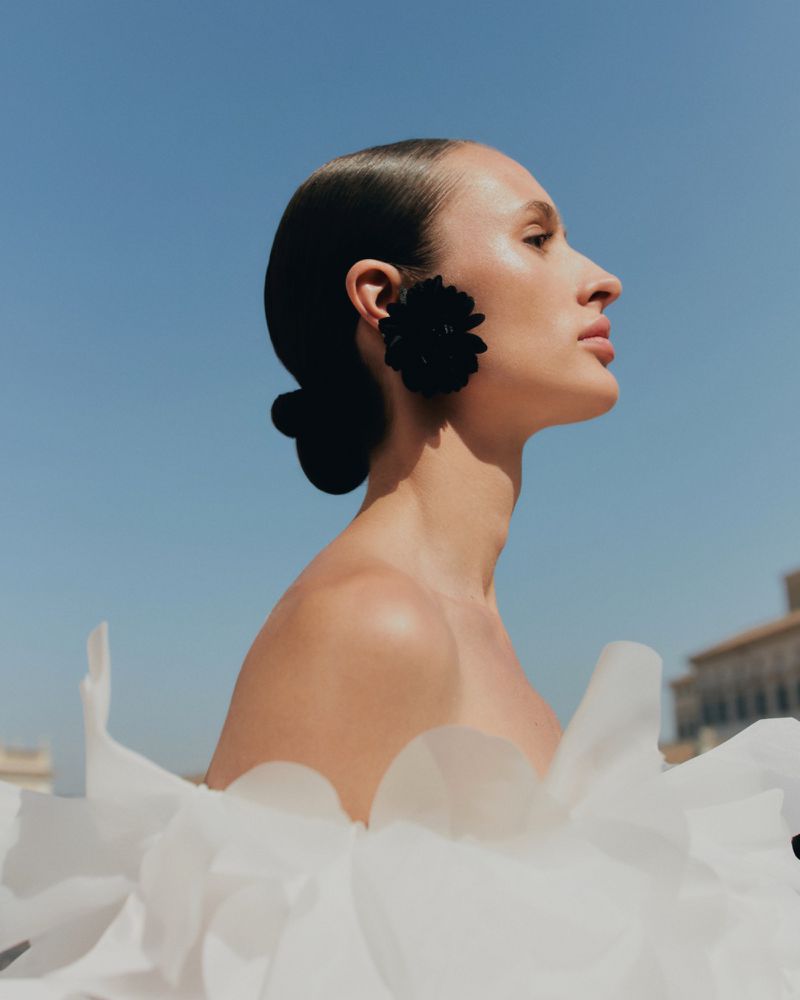 Elegant woman in profile wears oversized black floral earring and strapless white dress, posing against a bright blue sky on sunny day.