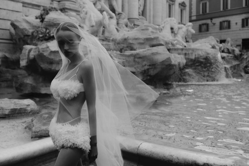 Monochrome image of a woman in a floral swimsuit and veil posing in front of the Trevi Fountain. Fashion editorial photography.