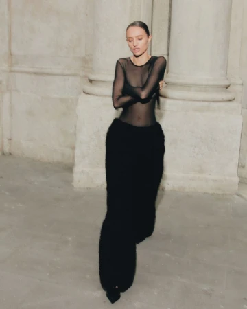 A woman in a black dress stands in a stone hallway with arches and columns, creating a vintage fashion photograph.