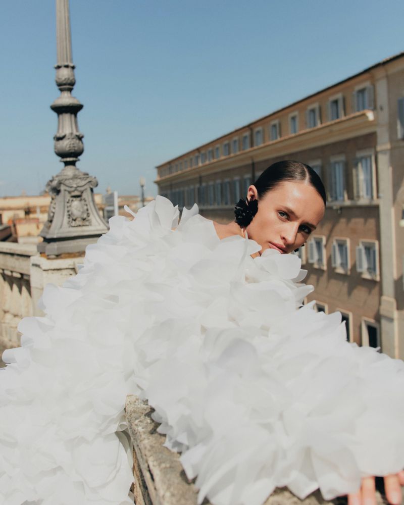 Woman in large white ruffled dress poses on balcony with city buildings in the background under clear blue sky, eye-level angle.