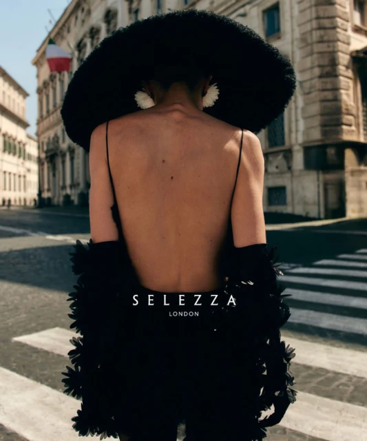 Fashion shot of a woman wearing a large black hat and dress with a feathered trim standing on a crosswalk in an urban environment.