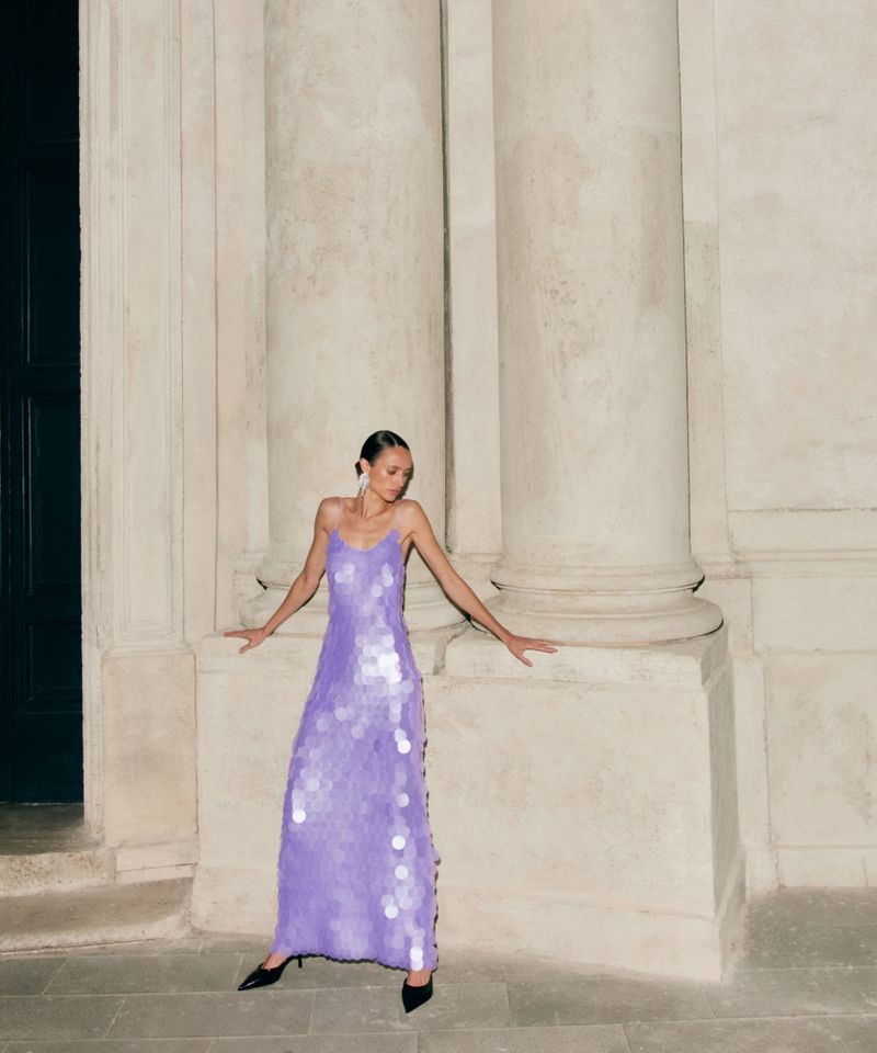 Elegant woman in a lilac sequin dress poses against architectural columns, fashion-forward style, with black heels and subtle jewelry.