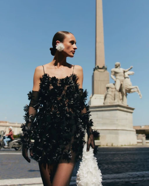 A woman models a black feathered dress on a cobblestone street with a tall obelisk and sculpture in the background under a blue sky.