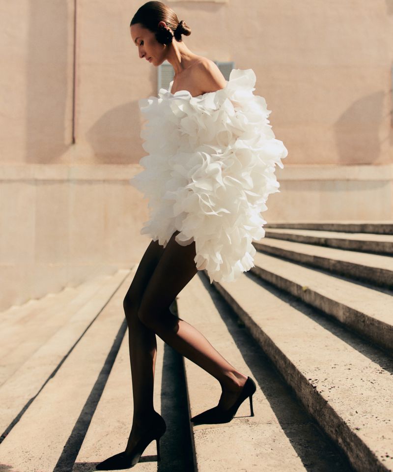 An adult woman is walking down steps wearing a white frilly dress, black stockings, and black heels in an outdoor urban environment.