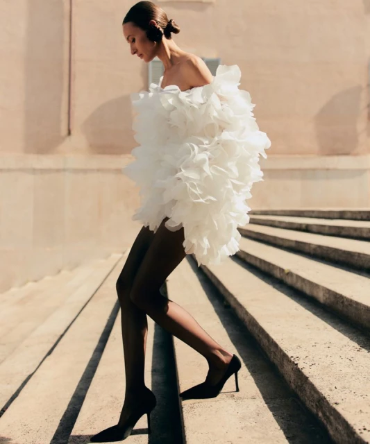 An adult woman is walking down steps wearing a white frilly dress, black stockings, and black heels in an outdoor urban environment.