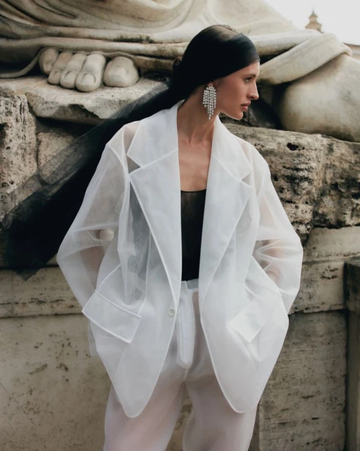 Elegant woman poses in a sheer white blazer and pants, accessorized with sparkling chandelier earrings, standing against a weathered stone statue.