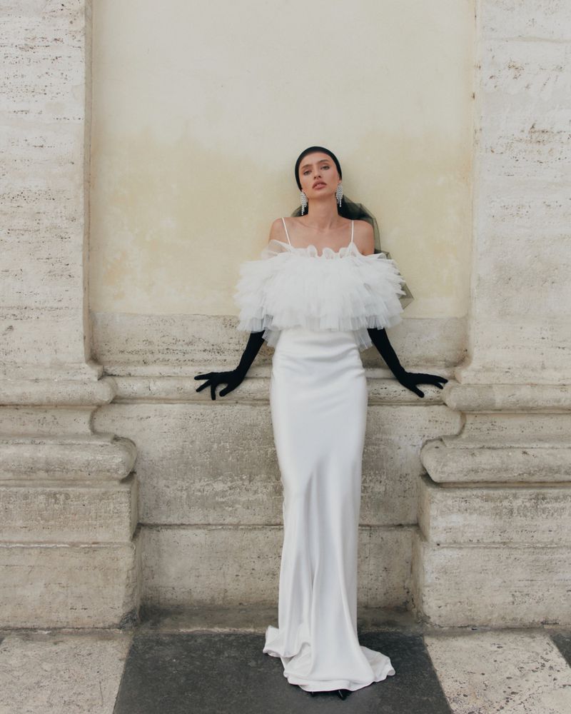 Woman in an elegant white gown with black gloves poses next to a fluted column in an outdoor setting, creating a striking visual.
