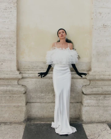 Woman in an elegant white gown with black gloves poses next to a fluted column in an outdoor setting, creating a striking visual.