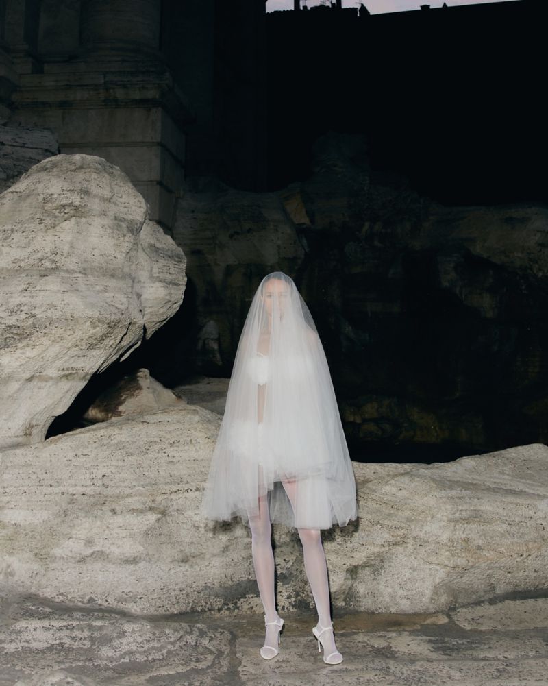 Woman in white dress and veil standing in front of an ancient stone wall at night with dark sky for fashion or conceptual photography.