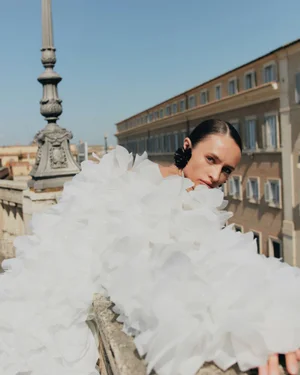 Model wearing a voluminous white ruffled dress leaning against a stone balustrade with a classical building and clear sky behind