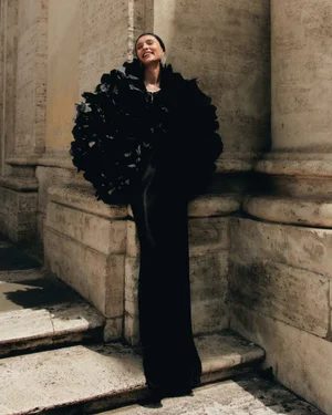 Woman in a voluminous black feathered dress leaning against classical stone architecture outdoors.
