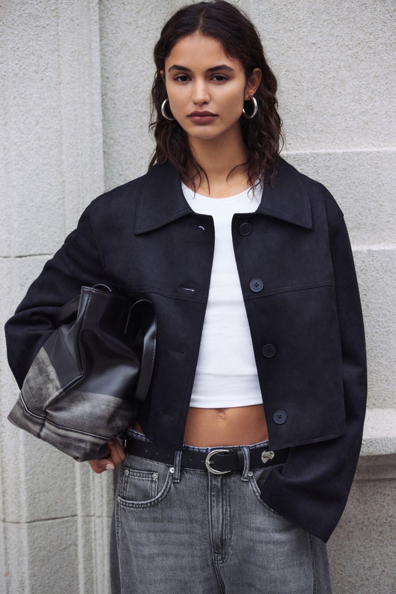 A woman in a cropped black jacket and gray jeans stands in front of a textured gray wall, holding a black bag, with natural lighting.