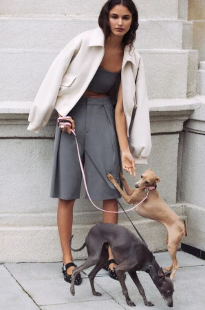 Woman in jacket and shorts with two Italian greyhounds on leashes in a city, walking up the stairs outside a stone building.
