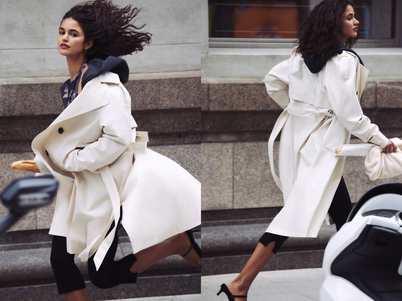 Adult female running with baguette and cream-colored trench coat and black leggings outdoors in an urban environment with neutral tones.