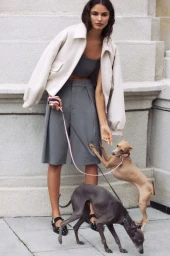 Model wearing a cream jacket and grey shorts holding leashes of two dogs standing on stone steps