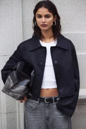 Model in black jacket and grey jeans holding a black handbag against a stone building exterior wall