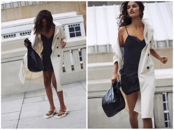 Model wearing a white trench coat over a black slip dress carrying a black handbag outdoors in an urban setting for Pull and Bear.