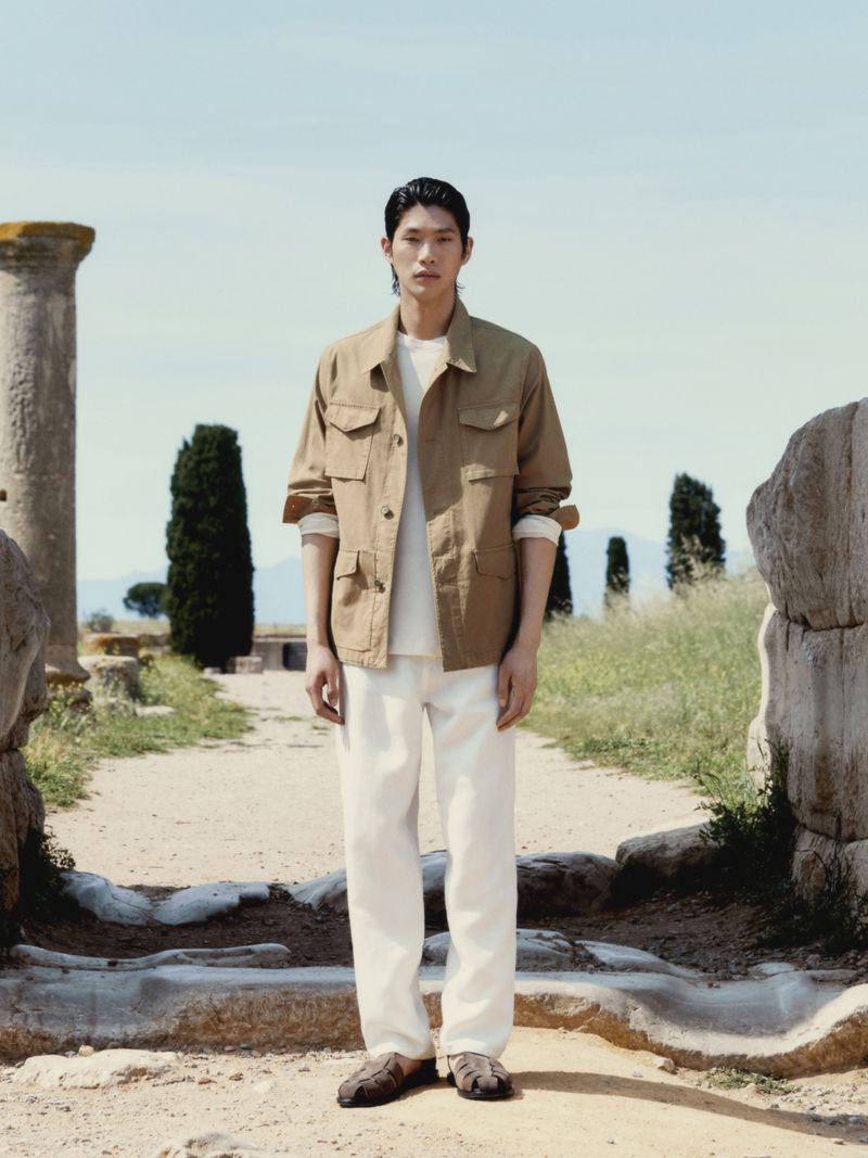 Adult male model stands in full shot on path with stone ruins wearing khaki chore jacket, white t-shirt, and white pants in outdoor setting.