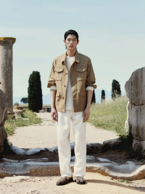 Adult male model stands in full shot on path with stone ruins wearing khaki chore jacket, white t-shirt, and white pants in outdoor setting.
