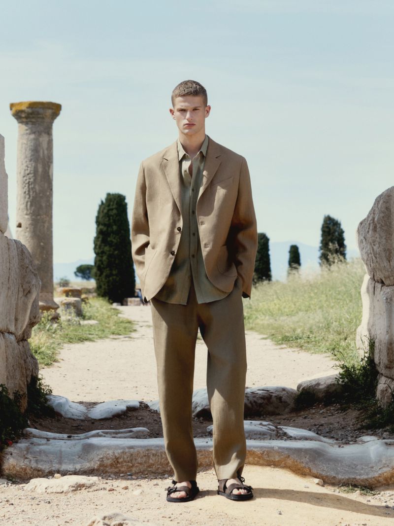 Male model in a tan linen suit and sandals standing outdoors among weathered stone ruins, with a blue sky backdrop. Fashion editorial.