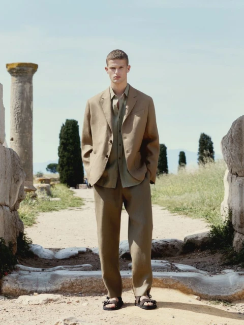 Male model in a tan linen suit and sandals standing outdoors among weathered stone ruins, with a blue sky backdrop. Fashion editorial.