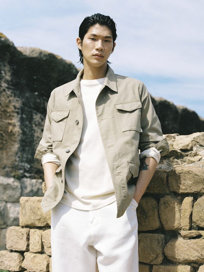 Asian male in a field jacket, white t-shirt, and white pants poses outside on a sunny day with a stone wall in the background.