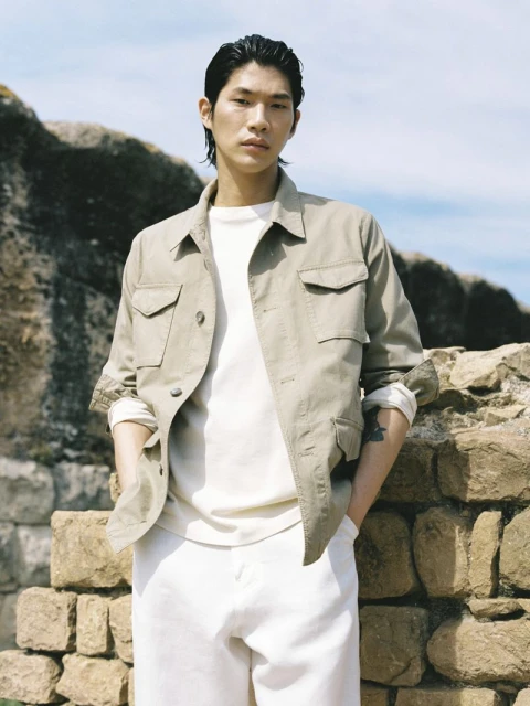 Asian male in a field jacket, white t-shirt, and white pants poses outside on a sunny day with a stone wall in the background.