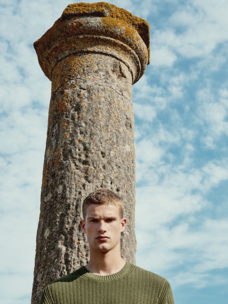 Portrait of a handsome man in an olive green sweater standing outdoors with an ancient stone column and a sky with clouds behind him.