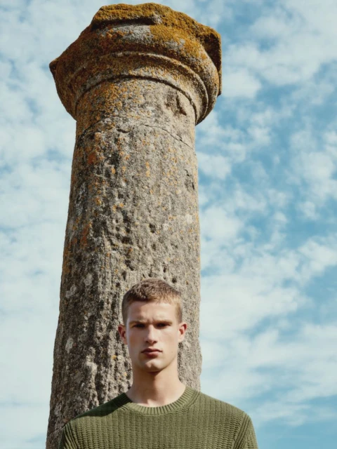 Portrait of a handsome man in an olive green sweater standing outdoors with an ancient stone column and a sky with clouds behind him.