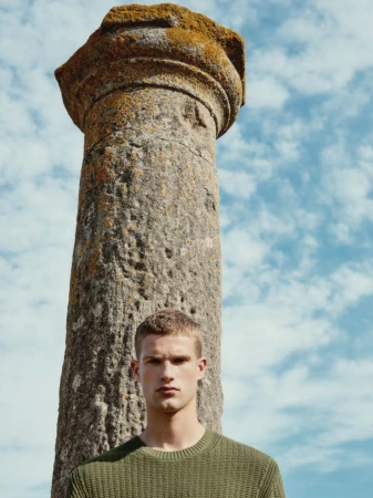 Portrait of a handsome man in an olive green sweater standing outdoors with an ancient stone column and a sky with clouds behind him.